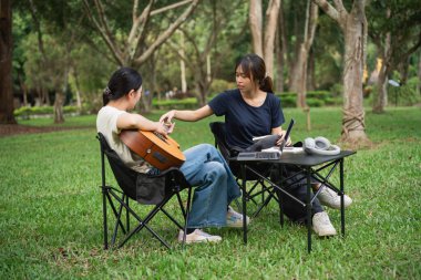 İki genç kadın parkta yaratıcı bir öğleden sonranın tadını çıkarırken, biri gitar çalarken diğeri müzik aletleri ve defterle masa üstünde takılıyor.