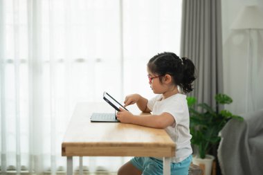 Gözlüklü genç kız evde tablet öğreniyor. Parlak, havadar bir odada. Etrafı kapalı bitkilerle çevrili.