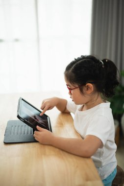 Evde tablet kullanan genç bir kız merak içinde, parlak ve rahat bir ortamda teknoloji yoluyla öğrenme ve dijital keşiflere girişiyor.