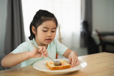 Evde kaşıkla kahvaltı eden genç bir kız, sıcacık bir mutfak ortamında tost yiyor, parlak doğal ışıkla yemeğine odaklanmış.