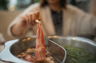 Yakın plan bir görüntü ince dilimlenmiş etin pişirme sürecini gösteriyor. Taze sebze ve aromatik et suyuyla dolu dumanı tüten sıcak bir tencereye batırılıyor..