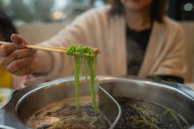 Bir kadın, erişteyi kaynayan sıcak bir tencerenin üzerinde ustalıkla tutuyor, sıcak bir restoranda lezzetli bir yemek sergiliyor, buhar ve canlı malzemelerle çevrili..
