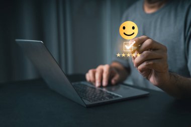 Yakın plan iş adamı eli, elinde gülen surat emojisi ve müşteri memnuniyeti ve çevrimiçi geri bildirimi temsil eden beş yıldızlı bir not sembolü tutarken dizüstü bilgisayarla etkileşime giriyor.