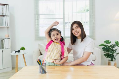 Sırt çantalı mutlu çocuk, çalışma saatleri boyunca gülümseyen anneyle poz veriyor. Parlak odada ebeveynlik bağı ve öğrenme ortamı gösteriyor.