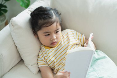 Genç bir çocuk rahat bir kanepede kitap okurken sakin bir anın tadını çıkarıyor. Parlak, davetkar bir oturma odasında. Bitkiler ve doğal ışıkla dolu.