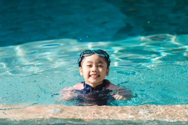 Mutlu çocuk havuzda yüzüyor, yüzme şapkası ve gözlüğü takıyor, yaz eğlencesinin tadını çıkarıyor, su sıçratıyor, parlak güneş ışığı yüzeye yansıyor.