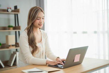 Modern ofis ortamında dizüstü bilgisayarla çalışan genç bir kadın, doğal ışıkla, görevlere odaklanmış ve verimliliği ve çağdaş çalışma alanı tasarımını gözler önüne sermiş.