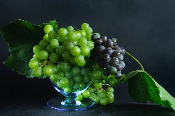 Clusters of black and green grapes with leaves in a glass bowl on a black shabby background in daylight. selective focus