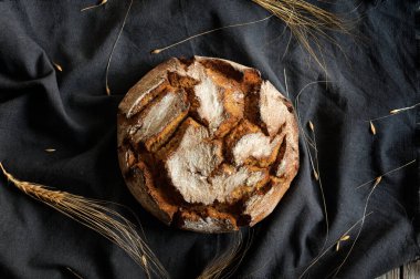 Tahıl ve ekmek kulaklı, koyu bir masa örtüsü üzerinde iştah açıcı yuvarlak bir ekmek somununun üst görüntüsü. Gün ışığı, seçici odak