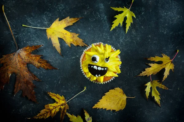 Halloween cupcake with a smiley face on a dark shabby background with autumn leaves. Overhead view, selective focus in daylight