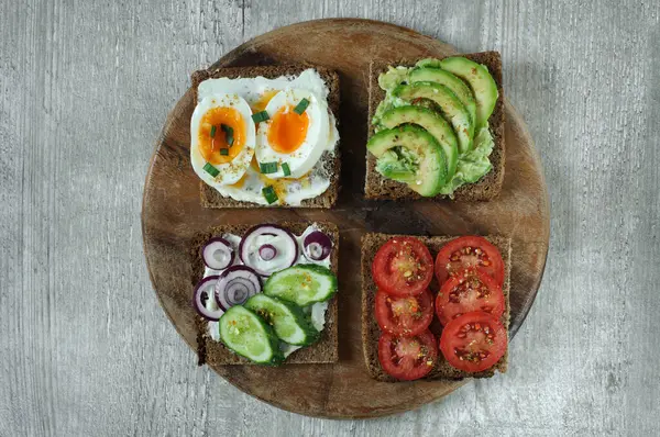 stock image Various sandwiches on a wooden board made of black rye whole grain bread with tomato, avocado, egg and cucumber. Top view