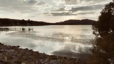 Sakin bir video günbatımında bir dağ gölü yakalıyor, sıcak renklerle boyanmış gökyüzü sakin suyu yansıtıyor, sakin ve nefes kesici bir doğal manzara yaratıyor.