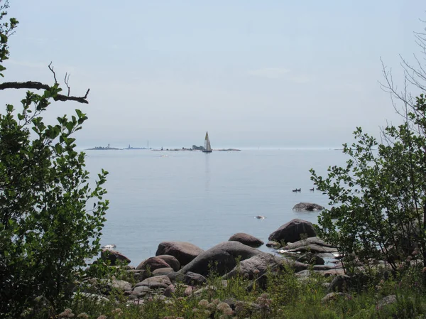 Sea view between trees and rocks with a sailboat in front of small rocky islets
