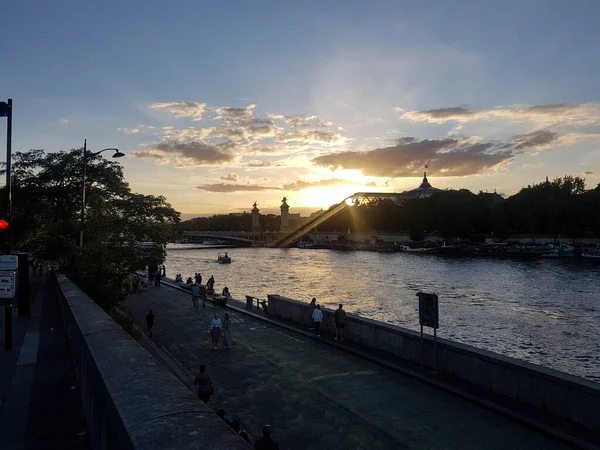 People walking on Seine bank in Paris with sun setting behind Grand Palais and Pont Alexandre III bridge