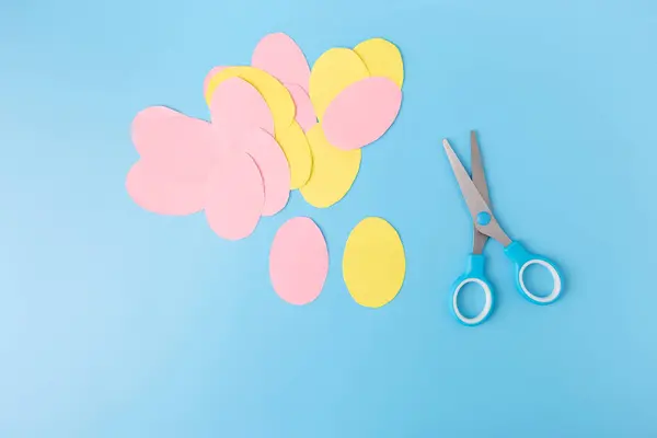 a pair of scissors and paper smooth round shapes eggs on a blue surface, a pastel colors, top view, step by step instruction, DIY, spring or Easter craft activity for kids,