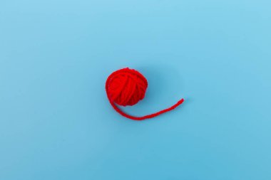 Sade mavi arka planda duran küçük kırmızı bir iplik yumağı. Bu resim, hem yaratıcı hem de fonksiyonel bir aktivite olan araçları ve malzemeleri temsil eder..