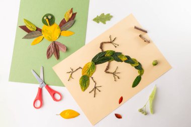 Creative leaf art on two pieces of paper, one green, one beige. Green: bird made from leaves, petals, googly eyes. Beige: lizard crafted with leaves, twigs. Natural materials scattered around. clipart