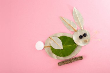 Pembe arka planda doğal malzemelerden yapılmış el yapımı bir tavşan. Yapraklar vücut ve kulakları, dal ve ağız için, yuvarlak malzemeler gözler için ve tüylü nesneler kuyruk olarak oluşturur. Yaratıcı, çekici, sanatsal.