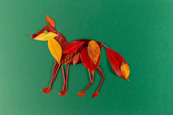 stock image Artistic fox crafted from red and yellow leaves against green backdrop. Leaf details depict body, head, legs, tail, ears, and nose. Creative natural material, innovative animal figure design.