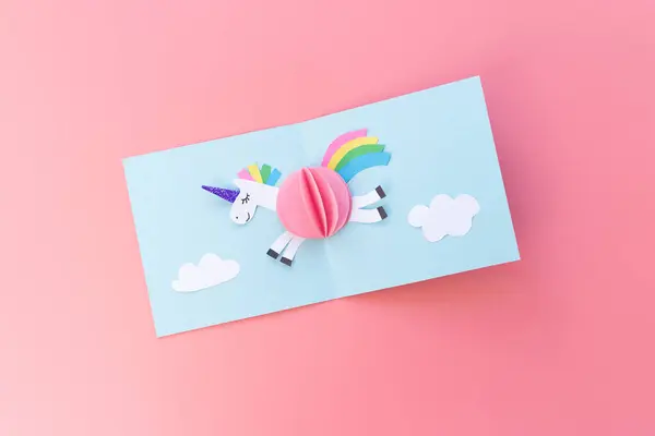 Gökyüzünde uçan tek boynuzlu bir atı tasvir eden pastel renkli kağıt kesiminin yukarıdan görünüşü, tam bir kart olarak tasarlandı. Tuhaf ve fantezi temalı projeler için mükemmel..