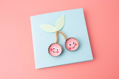 A pair of creatively crafted objects resembling cherries, made from bottle caps with pink centers and white dots, with green paper leaves. Playful and artistic craft, homemade postcard, DIY,  clipart