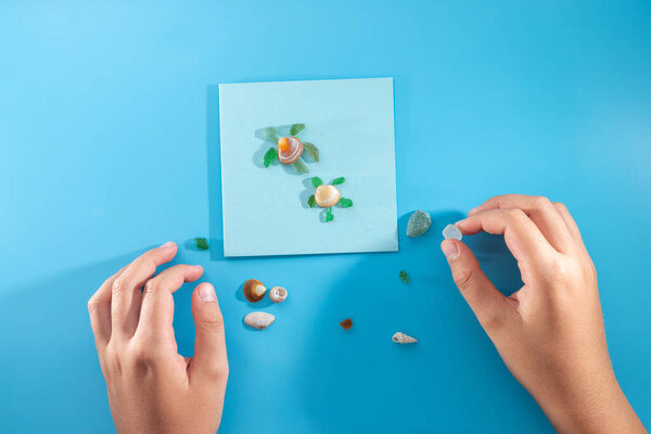 Creative turtle, child hands playing with sea shell, handmade turtle from recycled materials, smooth colorful sea glass pieces, cardboard, top view, blue background, tutorial, DIY, beach materials
