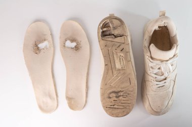 Eski, yıpranmış, tabanı kısmen ayrılmış ve tabanı eksik beyaz bir spor ayakkabı. Görünür kir, aşınma ve ayakkabıda delikler. Çıkarılmış taban ve taban tabanı hemen yanında.
