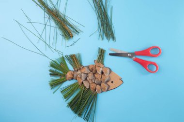 Çam kozalaklarından yapılmış balık şekilli tekneler ve mavi arka planda kırmızı makasla çam iğneleri. DIY proje konsepti. Eğitim veya zanaat tasarımı için düz konum