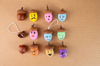 Boyanmamış meşe palamudlarının, boya paletlerinin ve bej arka planda fırçaların yanında emoji yüzleri olan el yapımı meşe palamutları. Kartlar için düz sonbahar sanatı fikri, DIY doğa sanatı projesi, sonbahar dekorasyonu