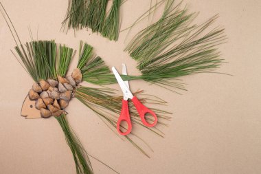 Çam kozalağı, çam iğnesi ve bej arka planda kırmızı makaslı karton kullanarak DIY balık gemisi. Yaratıcı süreçte adım, geri dönüşüm ve eğitim faaliyetleri, yukarıdan aşağıya görünüm