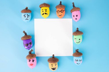 Mavi arka planda beyaz bir karenin etrafına dizilmiş boyanmış meşe palamudu koleksiyonu. Her meşe palamudunun farklı bir yüz ifadesi vardır ve farklı renklerde boyanmıştır.. 