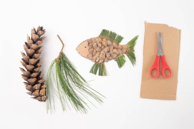 Bir çam kozalağının yanında oturan bir makas, çevre sanatı, tohum pullarından ya da braklardan ya da çam iğnelerinden yapılmış balıklar, kartondan kesilmiş hayvanlar, DIY, çocuklara özel ders, beyazlara tepeden bakmak.