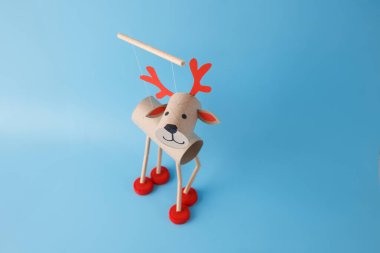 Karton tüplerden, kırmızı şişe kapaklarından ve kağıt detaylarından yapılmış ren geyiği kuklası. Mavi arka plan. Noel zanaatı ve tatil dekorasyonu. Çocuklar ve anaokulu için tuvalet kağıdı rulosu.