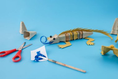 Açık mavi zemin üzerinde makas, boya fırçası ve mavi boya ile geri dönüşümlü karton iguana zanaat. Stüdyo fotoğrafçılığı ve fotokopi alanı. DIY ve çevre dostu tasarım, baskı konsepti.