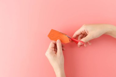 Hands cutting orange paper into crab claw shape with red scissors on a pink background. DIY craft process clipart