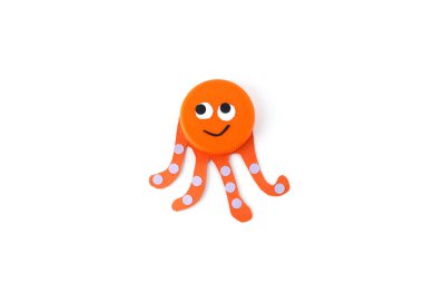 Orange plastic bottle cap transformed into an octopus face with paper tentacles and dotted details on a white . Minimalist flat lay composition. Craft and DIY recycling concept. homemade handicraft  clipart