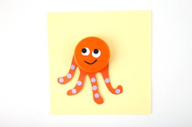 Turuncu plastik kapak kağıt dokunaçlı ahtapot yüzüne dönüştü ve sarı kare kağıtta noktalı detaylar var. Minimalist düz kompozisyon. Yapı ve DIY geri dönüşüm kavramı.