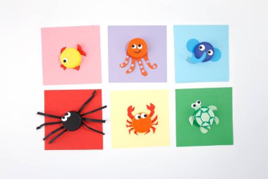 plastic bottle caps transformed into different samples of animals on color square paper. Minimalist flat lay composition on white background. DIY children summer activity and recycling concept. clipart