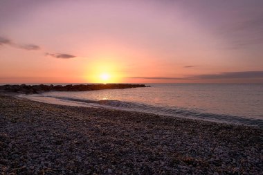Çakıl taşlı sahilde gündoğumu dalgalı suyla. Sonsuz arka plan, turuncu ve kırmızımsı renkler, boş plaj, küçük taşlar. Moncofar