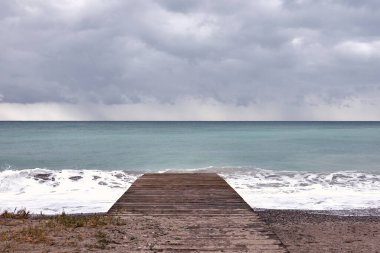 Wooden walkway on the shore of the beach. Storm clouds, swell, small stones, empty beach, grasses