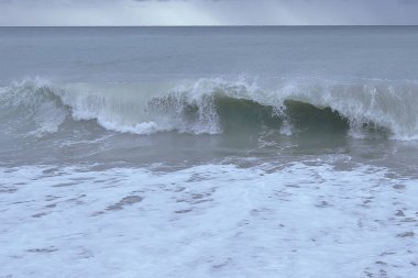 Küçük çakıl taşlı plajda dalgalar kıyıya vuruyor. Fırtına bulutları gökyüzü, turkuaz su, dalgalar,