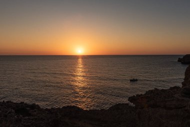 Şafak veya alacakaranlıkta sakin sularda süzülen bir tekne. Güneş ufkun ötesinde saklanıyor veya yükseliyor, turuncu ve mavi gökyüzü, bulutsuz, küçük tekne yelken açıyor, boş uzay..