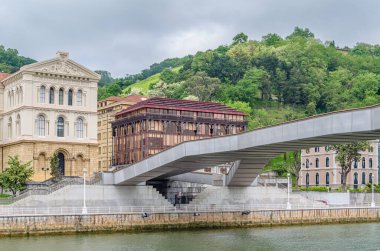 BILBAO, SPAIN - MAY 10, 2014: The Pedro Arrupe pedestrian walkway over the Nervion River in Bilbao, Basque Country, Spain, with the University of Deusto in the background clipart