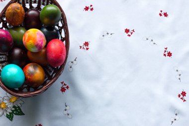 Beyaz masa örtüsü arkaplanındaki ahşap sepetteki renkli Paskalya yumurtalarının üst görüntüsü. Boşluğu kopyala.