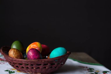 Siyah arka planda beyaz bir masa örtüsü üzerinde ahşap bir sepette renkli Paskalya yumurtaları. Boşluğu kopyala.