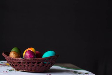 Siyah arka planda beyaz bir masa örtüsü üzerinde ahşap bir sepette renkli Paskalya yumurtaları. Boşluğu kopyala.