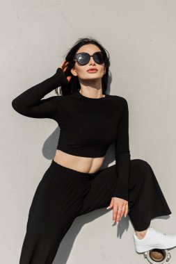 Moda siyah gözlüklü, beyaz spor ayakkabılı, siyah gözlüklü, şık bir genç kız gri bir duvarın yanında oturuyor ve poz veriyor.