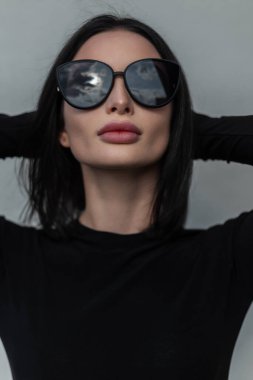 Modern şehir kadın portresi oldukça moda moda moda moda moda moda siyah elbiseli, havalı siyah güneş gözlüklü kız şehrin gri duvarının yanında poz veriyor.