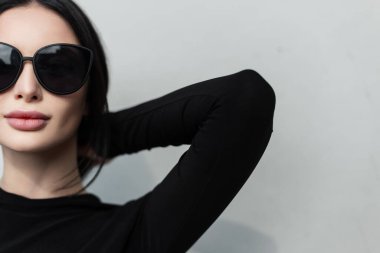 Şık, siyah, uzun kollu, gri bir arka plan yakınında poz veren siyah güneş gözlüklü güzel bir kadın modelinin modern kadın portresi. Yarım bir kadın yüzü