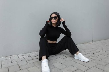Moda modasına uygun siyah güneş gözlüklü, beyaz spor ayakkabılı şık genç bir kadın modern gri bir binanın yanında sokakta oturuyor.